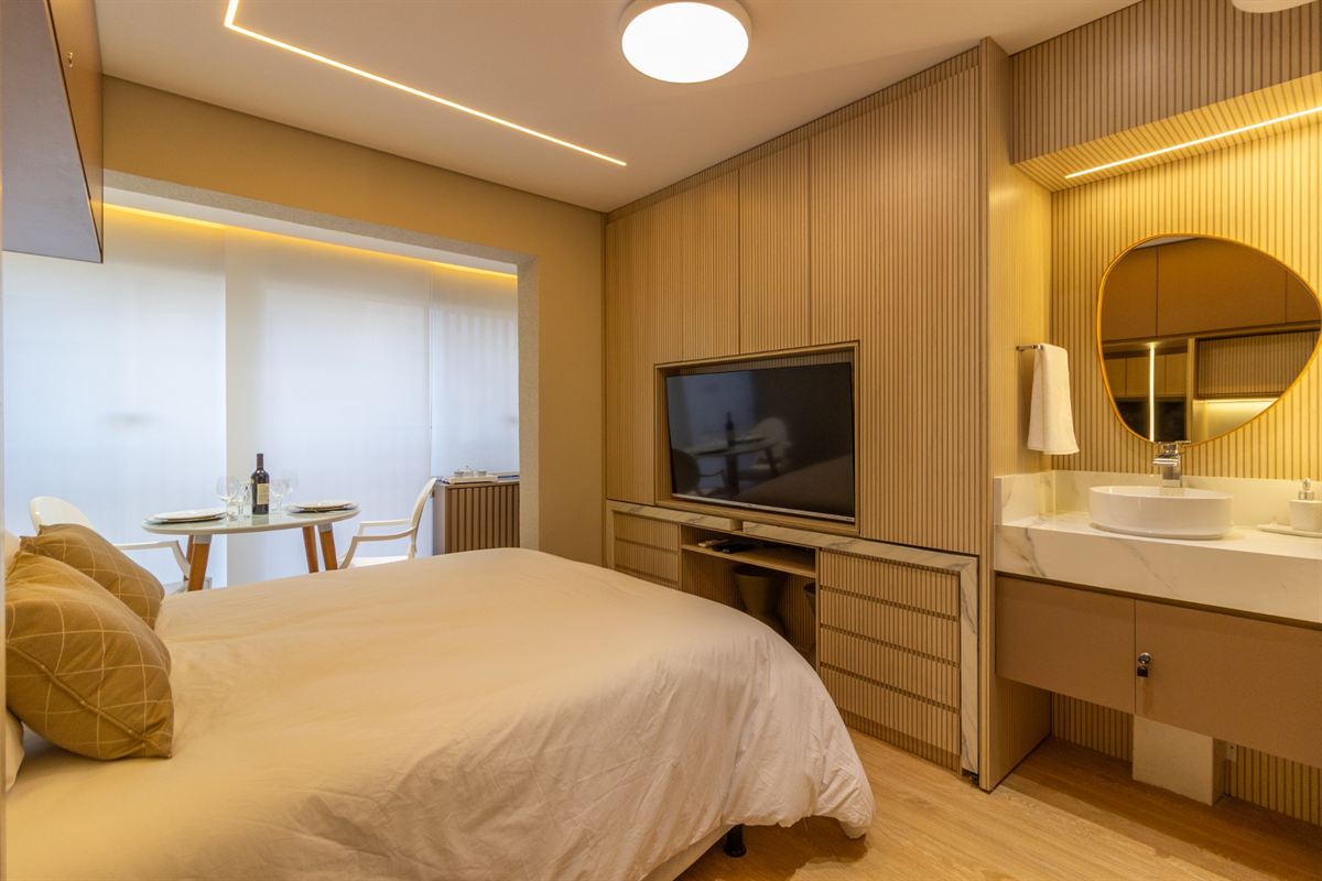 Quarto com cama de casal, TV em painel de madeira, bancada com cuba e mesa para duas pessoas
